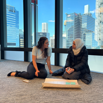 Two women sitting on the floor in conversation beside floor-to-ceiling windows overlooking a city skyline